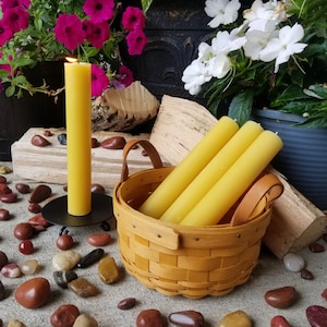 Op de afbeelding: Drie gele bijenwaskaarsen in een geweven mand met leren handvatten, omringd door rode en bruine stenen. Een enkele gele bijenwaskaars staat op een zwarte metalen kaarsenstandaard.