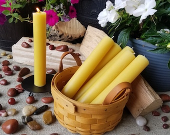 May include: A basket filled with multiple yellow beeswax candles, with one lit candle standing on a black pedestal. The basket is made of woven wood and has leather handles. The candles are arranged in a pile, with some of them sticking out of the basket. The scene is set on a gray surface with small red and brown stones scattered around.
