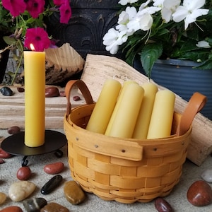 Puede incluir: Una cesta llena de velas de cera de abeja amarilla, con una sola vela de cera de abeja amarilla en un soporte de metal negro. La cesta está hecha de madera tejida con asas de cuero. Las velas están dispuestas en una pila, con algunas sobresaliendo de la cesta. También hay algunas piedras en el suelo alrededor de la cesta.