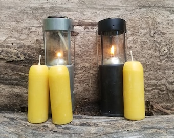 May include: Two metal candle lanterns with glass chimneys and lit candles, and two yellow beeswax candles on a wooden log.