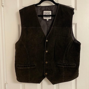 May include: A dark brown suede vest featuring a V-neckline, four metallic button closures down the front, and two horizontal welt pockets at the waist. The interior is lined with dark fabric and includes a label with the text BASIC.