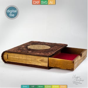 Puede incluir: Una caja de madera con forma de libro con un caj&oacute;n que se desliza hacia afuera. La parte superior de la caja tiene un dise&ntilde;o decorativo con un pentagrama y acentos florales. El caj&oacute;n est&aacute; forrado con fieltro rojo. La caja est&aacute; etiquetada como "Unique Creators" y tiene una escala de medici&oacute;n en pulgadas y mil&iacute;metros.