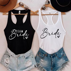 May include: Two tank tops with the text "Team Bride" and "Bride" in white script on a black and white tank top, respectively. The tank tops are hanging on a rack with hats on either side.