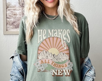 Peut inclure: T-shirt vert olive avec l'inscription "He Makes All Things New" dans un style rétro. Le motif comprend un soleil, des montagnes et des fleurs. Le t-shirt a des manches courtes et une coupe ample.