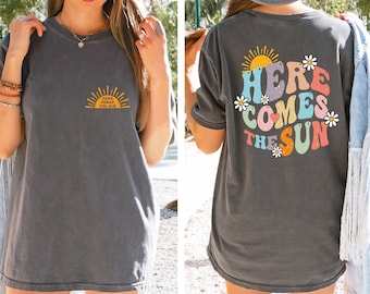 May include: A gray t-shirt with a graphic of a sun and the text "Here Comes The Sun" on the front and a larger graphic of the same design with the text "Here Comes The Sun" on the back.