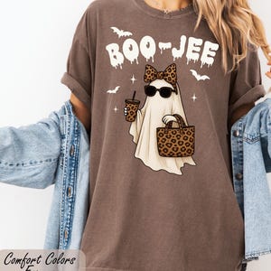 A woman wearing a brown t-shirt with a graphic of a ghost and leopard print accessories, including sunglasses and a handbag, standing in front of a white wall.