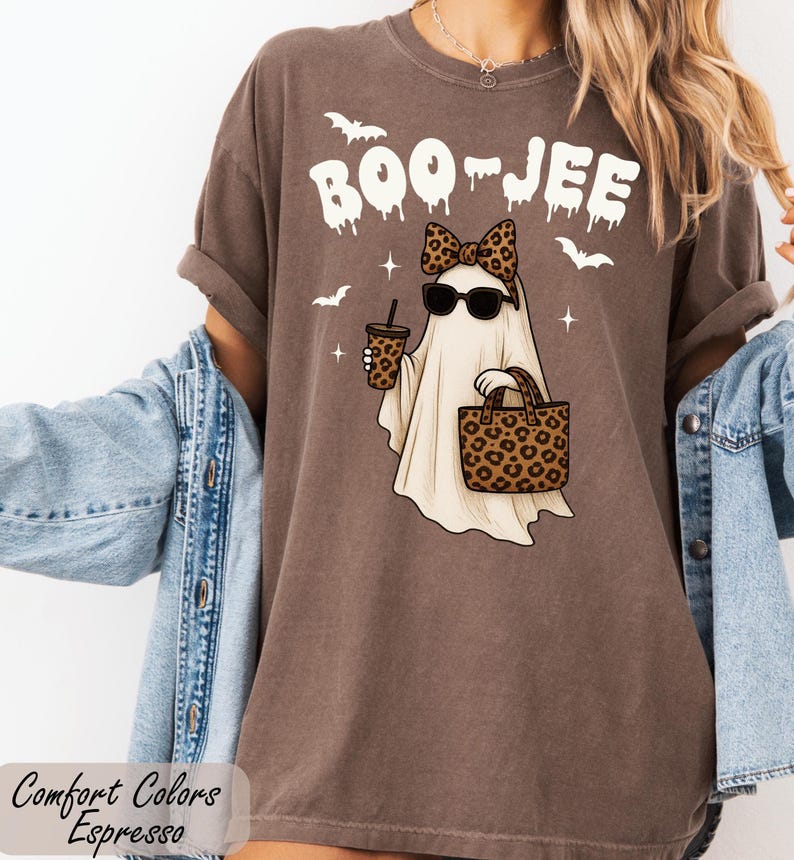 A woman wearing a brown t-shirt with a graphic of a ghost and leopard print accessories, including sunglasses and a handbag, standing in front of a white wall.