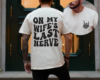 May include: A white t-shirt with black text that reads "On My Wife's Last Nerve". The text is in a retro font. The back of the shirt features a skeleton hand making the rock and roll hand gesture.