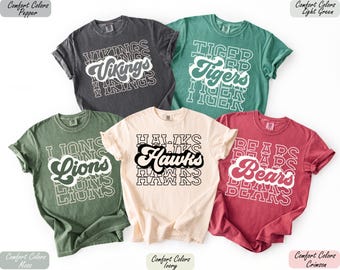May include: A collection of five t-shirts in various colors, each featuring a team name in a layered, retro font. The shirts are in shades of gray, green, ivory, red, and light green. The team names include "Vikings", "Tigers", "Lions", "Hawks", and "Bears".