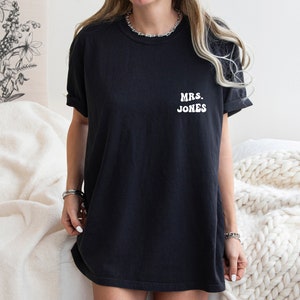 May include: Black t-shirt with the text "MRS. JONES" in white, a casual and comfortable style. The shirt is a dark color, likely cotton, and has short sleeves. The text is centered on the chest.