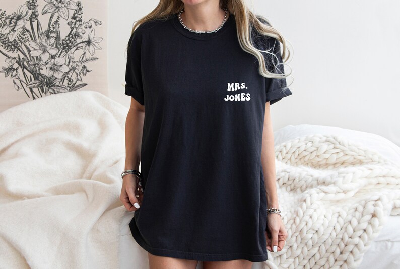 May include: Black t-shirt with the text "MRS. JONES" in white, a casual and comfortable style. The shirt is a dark color, likely cotton, and has short sleeves. The text is centered on the chest.