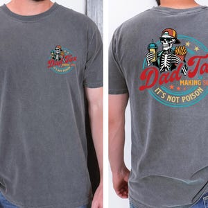 May include: Charcoal gray t-shirt with a graphic on the front and back. The front graphic is a skeleton with a drink and fries, and the text "Dad Tax". The back graphic is a skeleton with a drink and fries, and the text "Dad Tax Making Sure It's Not Poison".