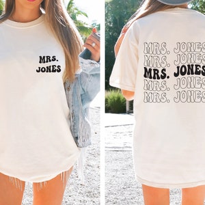 May include: Cream-colored t-shirt with the text "MRS. JONES" in a retro font on the front and back. The back of the shirt repeats the text multiple times. A casual, everyday wear item.