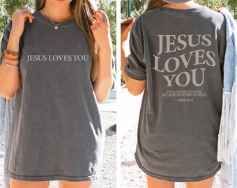 May include: A gray oversized t-shirt with the text "JESUS LOVES YOU" on the front and "JESUS LOVES YOU WE LOVE EACH OTHER BECAUSE HE LOVED US FIRST 1 JOHN 4:19" on the back.