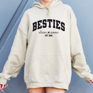 May include: A beige hoodie with the text "BESTIES" in black, with the names "alice" and "anna" and the date "EST. 2020" underneath.