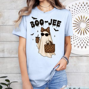 A woman wearing a light blue t-shirt with a graphic design of a ghost holding a drink and sunglasses, along with the text "BOO JEE".