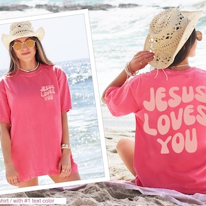 a woman sitting on the beach wearing a pink jesus loves you t - shirt