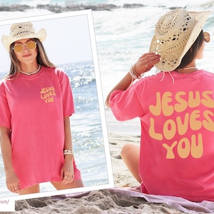 a woman sitting on the beach wearing a pink jesus loves you t - shirt