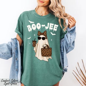 A woman wearing a green t-shirt with a graphic of a ghost and sunglasses, along with a denim jacket and a leopard print purse, posing against a white background.