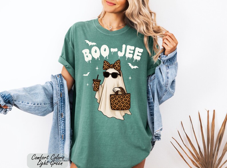 A woman wearing a green t-shirt with a graphic of a ghost and sunglasses, along with a denim jacket and a leopard print purse, posing against a white background.