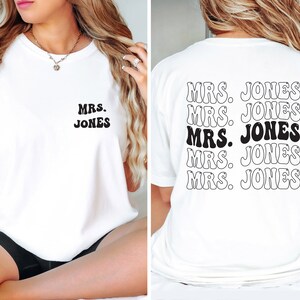 May include: White t-shirt with the text "MRS. JONES" in a retro font on the front and back. The back of the shirt repeats the text multiple times. The text is black and white.