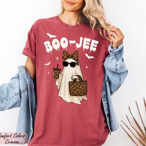 A woman wearing a red t-shirt with a graphic of a ghost and sunglasses, along with a denim jacket, posing against a white background.