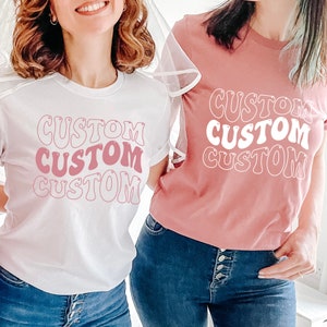 Op de afbeelding: Twee witte T-shirts met het woord "CUSTOM" bedrukt in roze en witte retro-lettertype. De shirts worden gedragen door twee mensen, een draagt een wit shirt en de ander een roze shirt.
