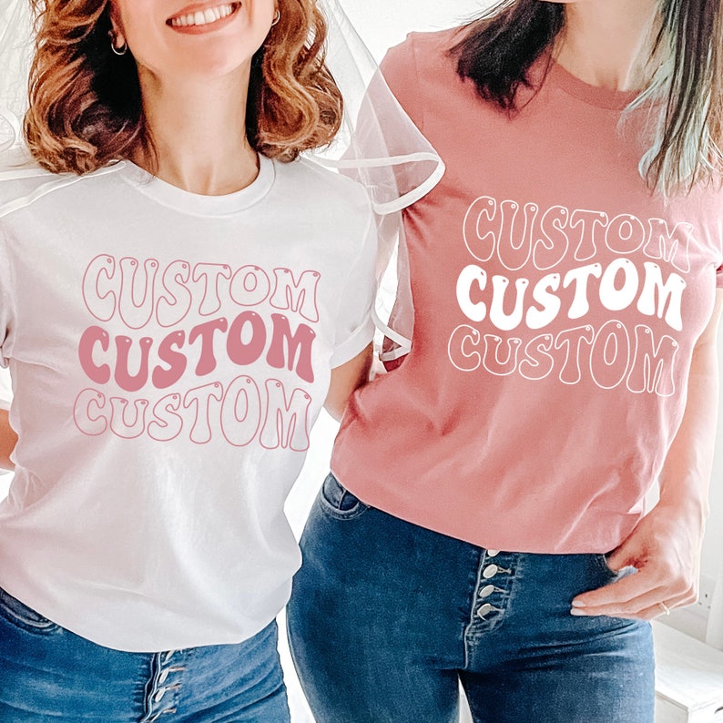 Op de afbeelding: Twee witte T-shirts met het woord "CUSTOM" bedrukt in roze en witte retro-lettertype. De shirts worden gedragen door twee mensen, een draagt een wit shirt en de ander een roze shirt.