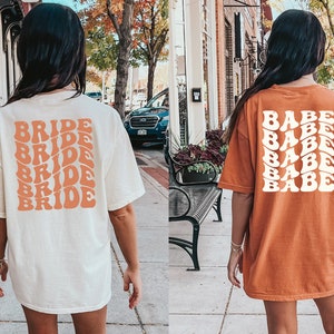 May include: Two women wearing oversized t-shirts. The white shirt has the word "BRIDE" repeated five times in a retro font in peach. The orange shirt has the word "BABE" repeated five times in a retro font in white.