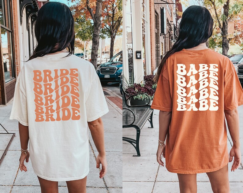 May include: Two women wearing oversized t-shirts. The white shirt has the word "BRIDE" repeated five times in a retro font in peach. The orange shirt has the word "BABE" repeated five times in a retro font in white.
