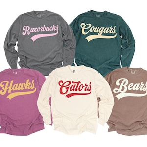 May include: Five long-sleeved shirts in various colors, each displaying a team name in a script font. The shirts are in shades of gray, green, purple, cream, and brown. The team names include "Razorbacks," "Cougars," "Hawks," "Gators," and "Bears."