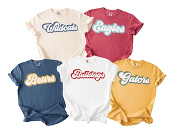 May include: Five crew neck t-shirts in various colors, each with a team name in a retro font. The shirts are cream, red, navy blue, white, and yellow. The team names are Wildcats, Eagles, Bears, Bulldogs, and Gators.