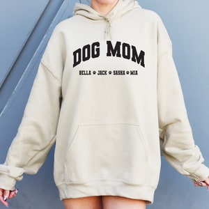 K&ouml;nnte beinhalten: Ein beiger Oversize-Hoodie mit dem Text "DOG MOM" in Schwarz und den Namen "BELLA, JACK, SASHA, MIA" in Schwarz darunter.