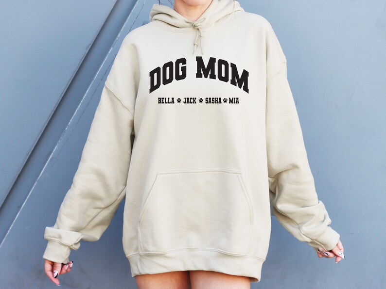 K&ouml;nnte beinhalten: Ein beiger Oversize-Hoodie mit dem Text "DOG MOM" in Schwarz und den Namen "BELLA, JACK, SASHA, MIA" in Schwarz darunter.