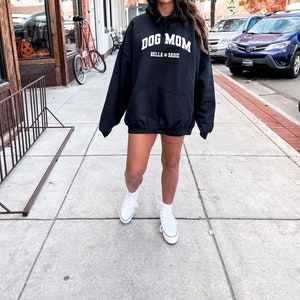 K&ouml;nnte beinhalten: Ein schwarzer Kapuzenpullover mit dem Text "Dog Mom" in wei&szlig;en Buchstaben. Der Text "Bella & Sadie" ist ebenfalls in wei&szlig;en Buchstaben unter dem Haupttext. Der Pullover wird von einer Person getragen, die wei&szlig;e Turnschuhe tr&auml;gt.