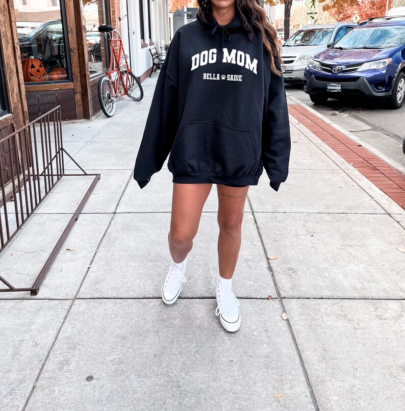 K&ouml;nnte beinhalten: Ein schwarzer Kapuzenpullover mit dem Text "Dog Mom" in wei&szlig;en Buchstaben. Der Text "Bella & Sadie" ist ebenfalls in wei&szlig;en Buchstaben unter dem Haupttext. Der Pullover wird von einer Person getragen, die wei&szlig;e Turnschuhe tr&auml;gt.