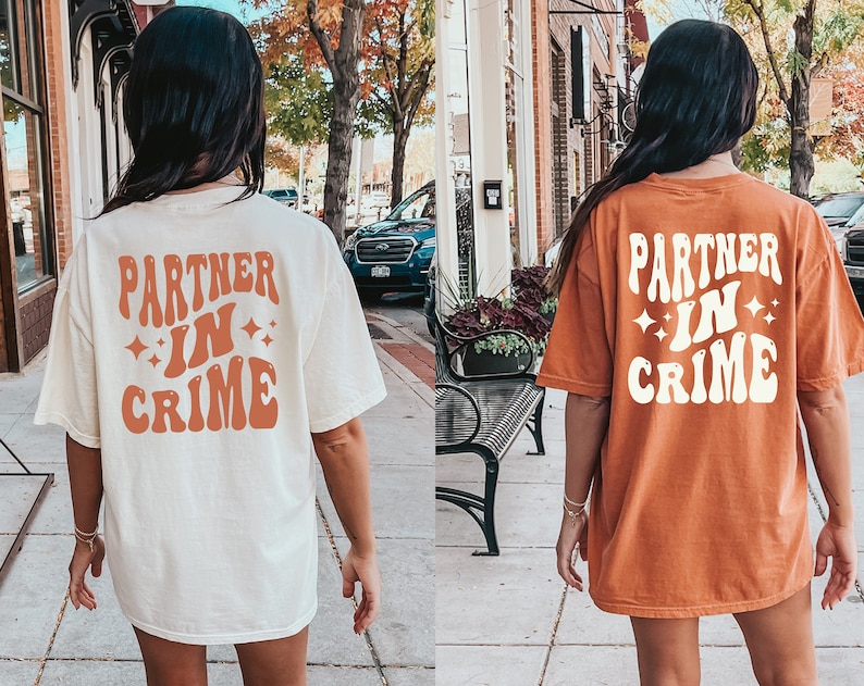 May include: Two people wearing matching t-shirts with the text "Partner in Crime" in a retro font. The shirts are white and orange.