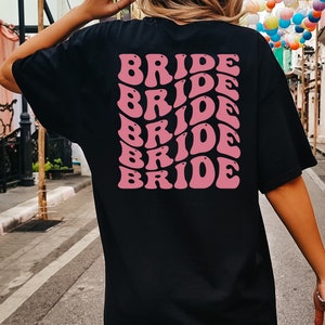 May include: Black t-shirt with the word "BRIDE" repeated in pink, retro-style lettering on the back.