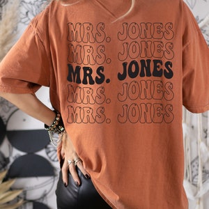 May include: Burnt orange t-shirt with the text "MRS. JONES" repeated in a stacked design on the back. The text is in a retro font with a black outline. The shirt has a relaxed fit and short sleeves.