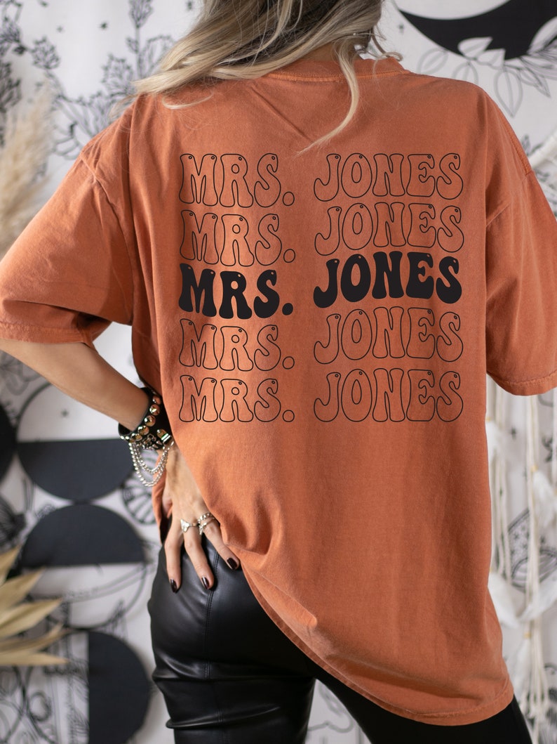 May include: Burnt orange t-shirt with the text "MRS. JONES" repeated in a stacked design on the back. The text is in a retro font with a black outline. The shirt has a relaxed fit and short sleeves.