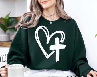 May include: A dark green sweatshirt featuring a white heart and cross design. The graphic is a simple, hand-drawn style. The sweatshirt is a crew neck style and appears to be made of a soft material.