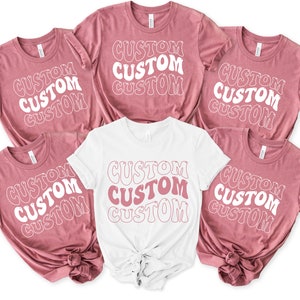 Op de afbeelding: Vijf T-shirts met het woord "CUSTOM" dat vier keer wordt herhaald in een retro-lettertype. De shirts zijn in twee kleuren: stoffig roze en wit.