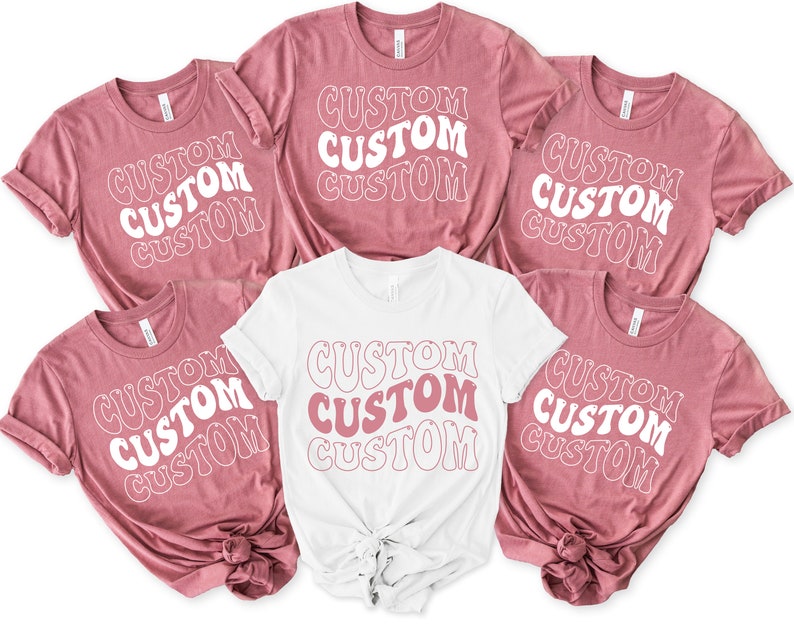Op de afbeelding: Vijf T-shirts met het woord "CUSTOM" dat vier keer wordt herhaald in een retro-lettertype. De shirts zijn in twee kleuren: stoffig roze en wit.