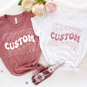 Op de afbeelding: Twee T-shirts met het woord "CUSTOM" geschreven in een retro-lettertype. De shirts zijn in verschillende kleuren, een is roze en de andere is wit. De shirts liggen op een wit, pluche oppervlak met een fles mousserende wijn.