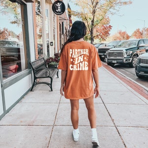 May include: A woman wearing a burnt orange t-shirt with the text "Partner in Crime" in white. She is walking down a sidewalk in front of a brick building.