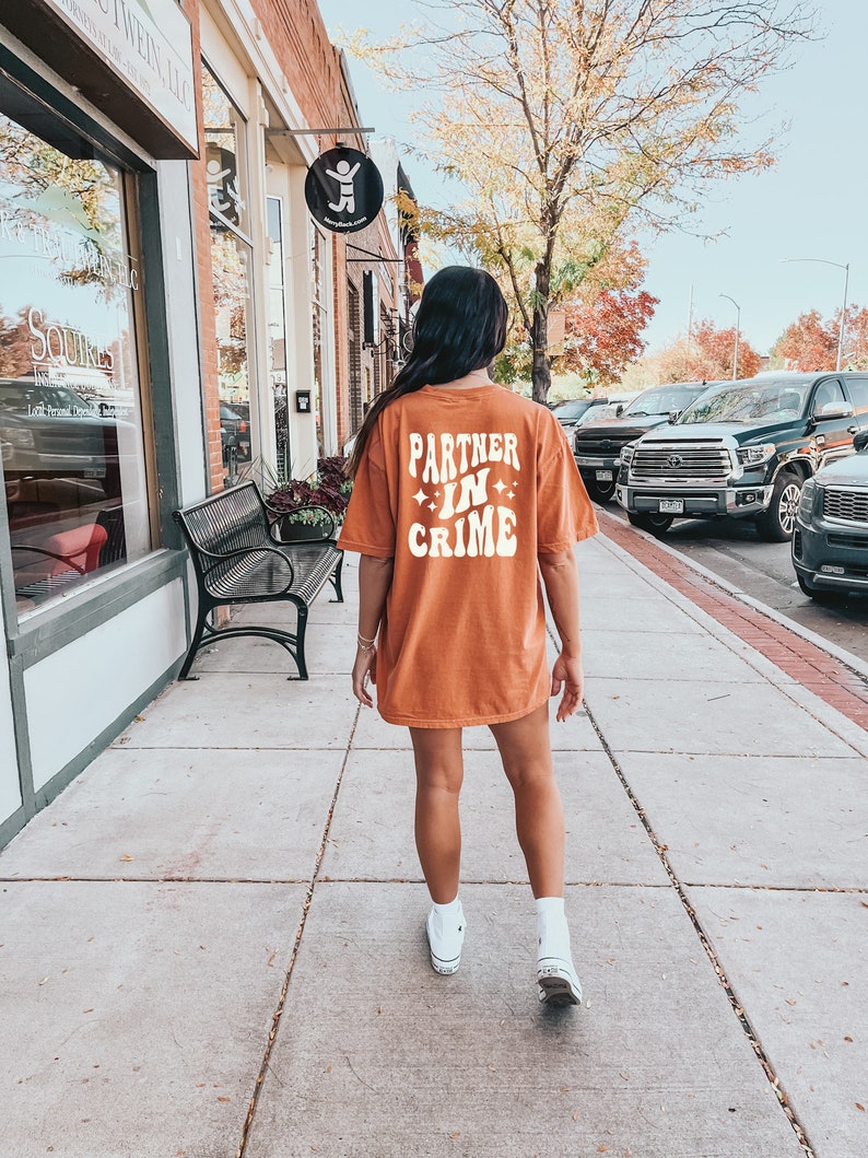 May include: A woman wearing a burnt orange t-shirt with the text "Partner in Crime" in white. She is walking down a sidewalk in front of a brick building.