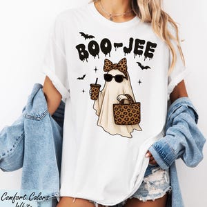 A woman wearing a white t-shirt with a graphic of a ghost and sunglasses, along with a denim jacket and denim shorts.