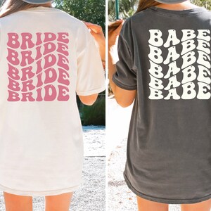 May include: Two women wearing oversized t-shirts. The white t-shirt has the word "BRIDE" repeated in pink, retro-style lettering. The gray t-shirt has the word "BABE" repeated in white, retro-style lettering.