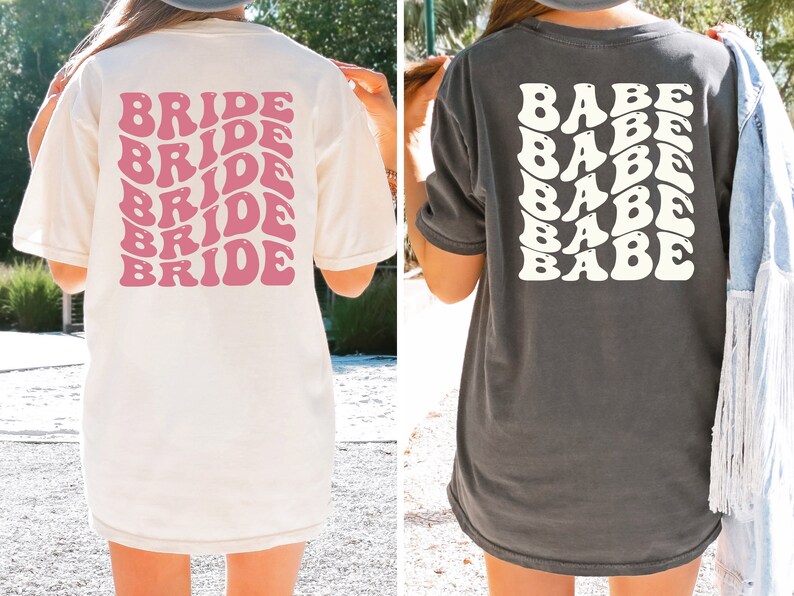May include: Two women wearing oversized t-shirts. The white t-shirt has the word "BRIDE" repeated in pink, retro-style lettering. The gray t-shirt has the word "BABE" repeated in white, retro-style lettering.