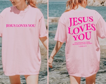 May include: Pink t-shirt with the text "JESUS LOVES YOU" in pink on the front and the same text in pink with the additional text "WE LOVE EACH OTHER BECAUSE HE LOVED US FIRST. 1 JOHN 4:19" on the back.